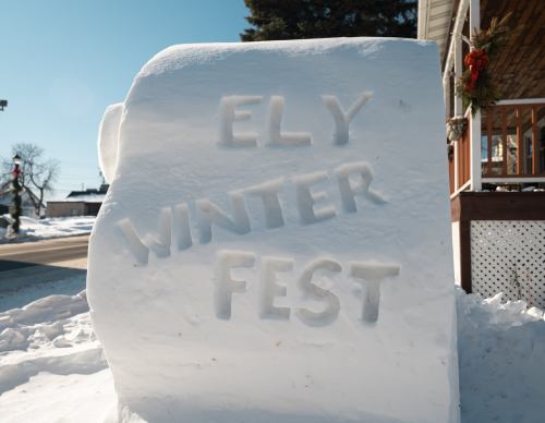 A large snow block with the words "ELY WINTER FEST" carved into it, surrounded by snow and a clear blue sky.