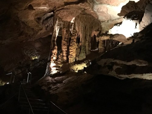 Stalactites and stalagmites illuminated in a dark cave, with a staircase leading into the cavern.