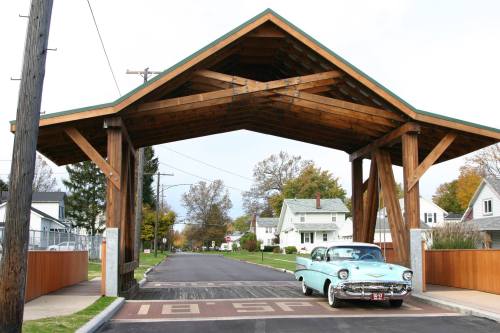 A vintage car drives under a wooden archway on a quiet street lined with houses and trees.