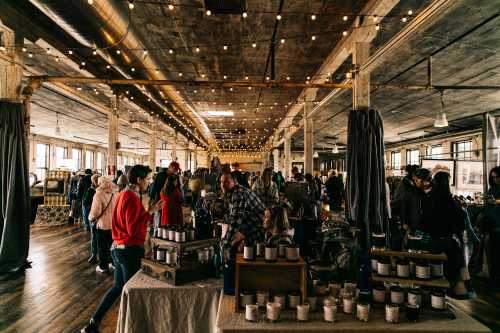 A bustling indoor market with people browsing stalls, string lights overhead, and various products displayed on tables.