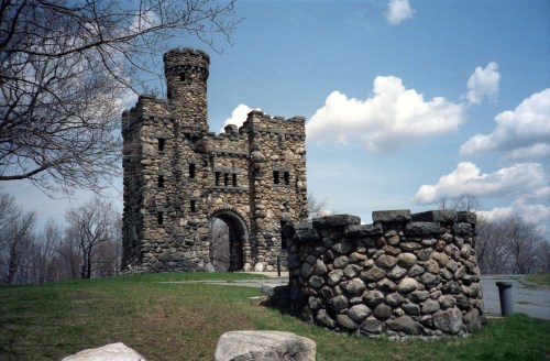 A stone castle with a tower and archway, surrounded by grass and trees under a partly cloudy sky.