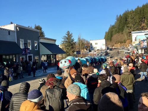 A crowd gathers on a street, watching a colorful parade float resembling a fish, with shops and trees in the background.
