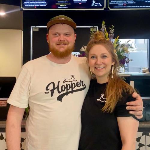 A smiling man and woman stand together in a restaurant, wearing matching branded shirts, with a menu in the background.