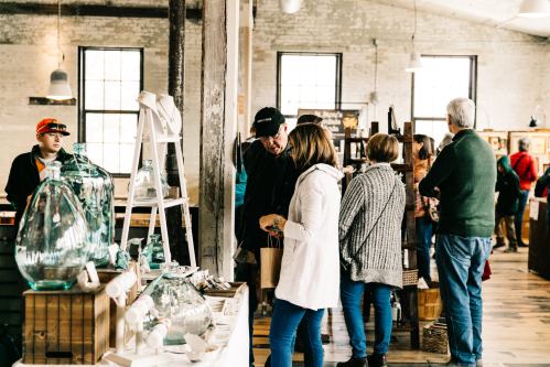 A busy market scene with people browsing handmade goods and chatting in a bright, rustic space.