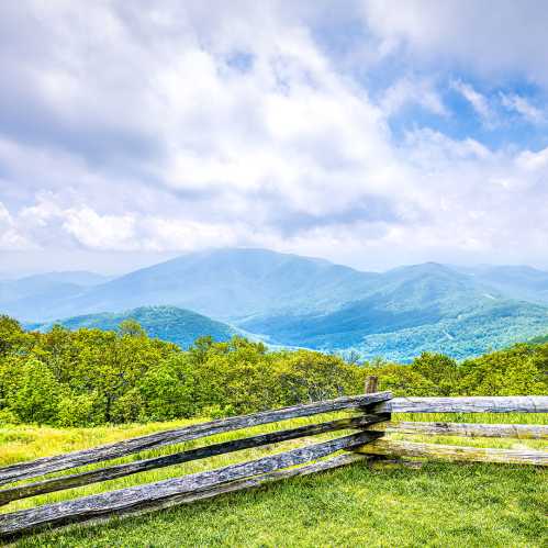 Scenic view of rolling green mountains under a cloudy sky, with a wooden fence in the foreground.