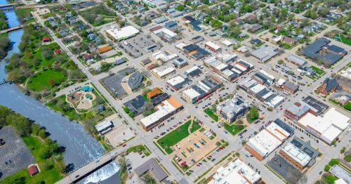 Aerial view of a small town with a river, parks, and a grid of streets lined with buildings and parking areas.