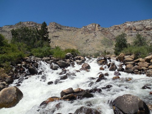 A rushing river flows over rocks, surrounded by greenery and steep rocky cliffs under a clear blue sky.
