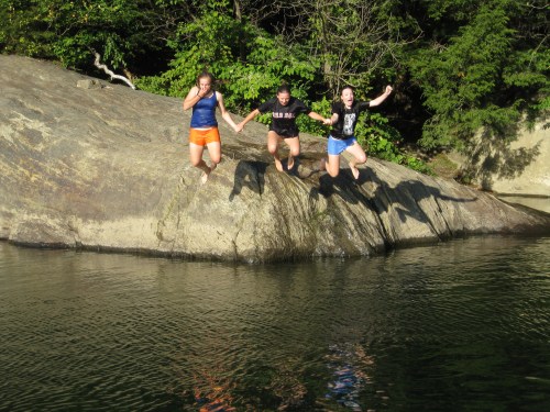 Three girls jump off a rock into a lake, surrounded by trees on a sunny day.
