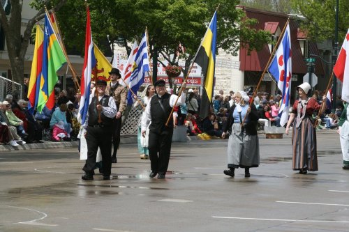 A parade featuring people in traditional costumes carrying flags, with a crowd watching in the background.
