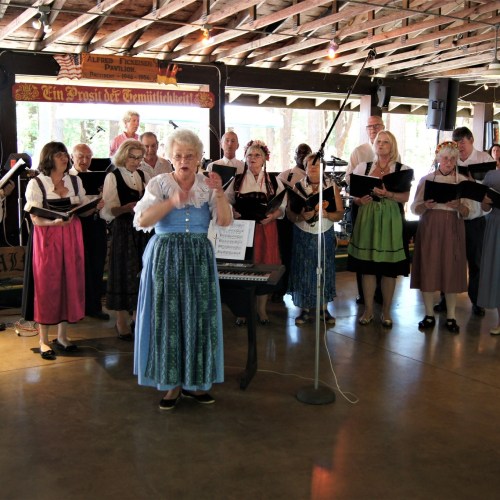 A choir in traditional attire performs on stage, with a conductor leading and musicians accompanying.