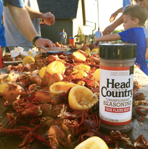A table covered with crawfish, lemons, and Head Country seasoning, with people enjoying a seafood feast outdoors.