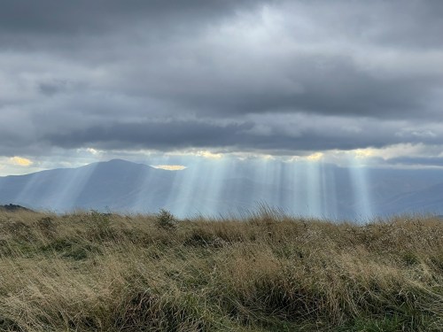 Sunlight streams through clouds over a grassy landscape, illuminating distant mountains in a dramatic sky.