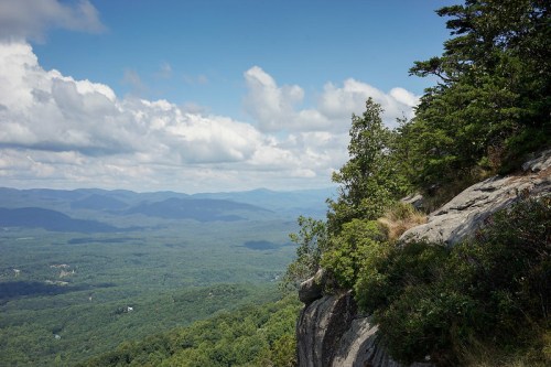 A scenic view from a rocky outcrop, overlooking lush green mountains and a cloudy blue sky.