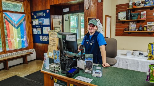 A smiling employee stands at a reception desk with a computer, surrounded by informational displays and a colorful flag.