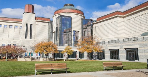 Modern building with large glass windows, surrounded by trees and benches on a sunny day.