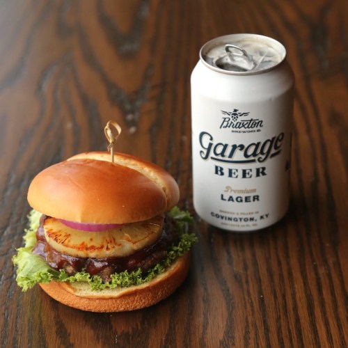A burger with lettuce, grilled pineapple, and onion, next to a can of Garage Beer on a wooden table.