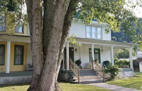 A charming two-story house with a porch, surrounded by greenery and a large tree in the foreground.