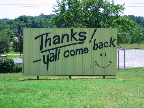 A large green sign with the text "Thanks! - y'all come back" and a smiling face, set against a grassy background.