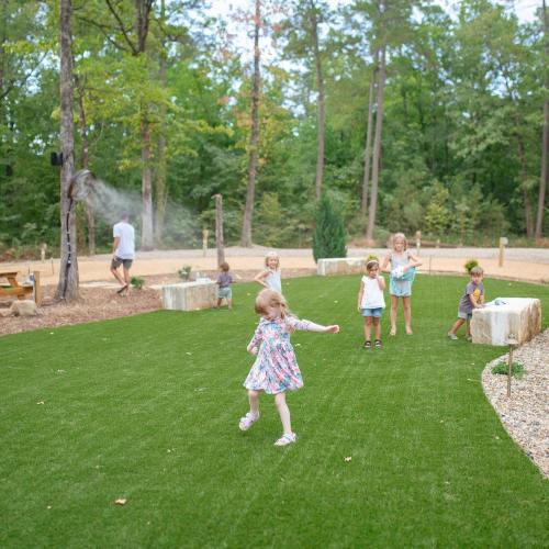 Children playing on a grassy area in a wooded park, with a few adults in the background.