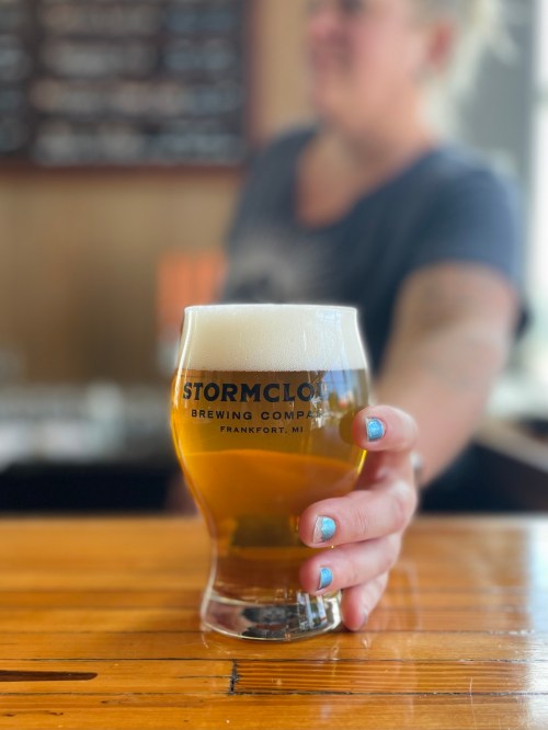 A person holds a glass of beer with a frothy head, featuring the Stormcloud Brewing Company logo, on a wooden bar.