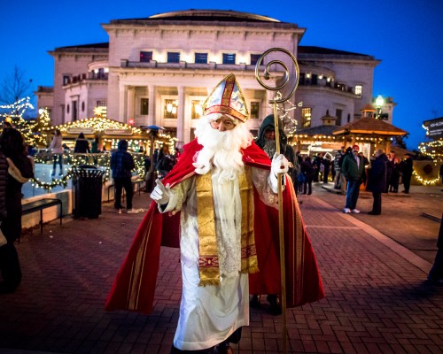 A figure in a red and white robe with a staff, resembling Santa, walks through a festive, illuminated outdoor market.