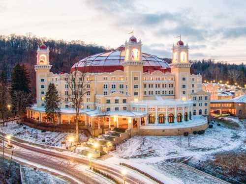 A grand hotel with a domed roof, surrounded by trees and snow, illuminated at dusk.