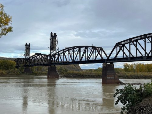 A steel bridge spans a river, with cloudy skies and autumn foliage in the background.