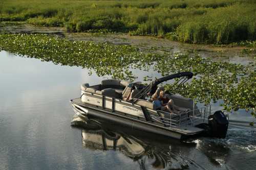 A pontoon boat glides through a calm waterway surrounded by lush greenery and lily pads. Two people enjoy the ride.