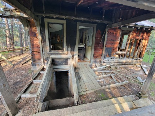 An abandoned, weathered cabin with a broken porch and darkened interior, surrounded by trees and overgrown vegetation.