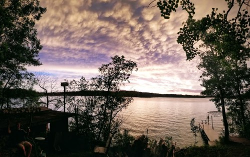 A serene lakeside view at sunset, with colorful clouds and trees framing the water's edge.