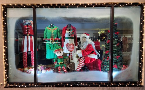 A festive window display featuring Santa, an elf, and holiday outfits, adorned with lights and decorations.