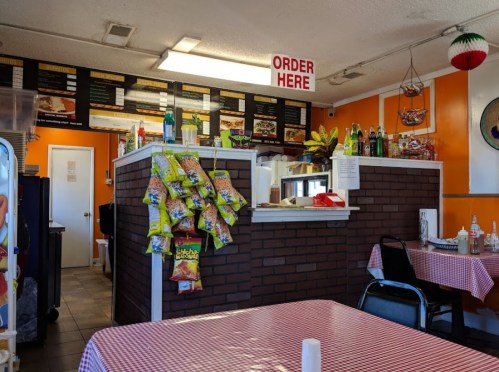 Interior of a casual eatery with a counter, menu board, snack bags on the wall, and checkered tablecloths.