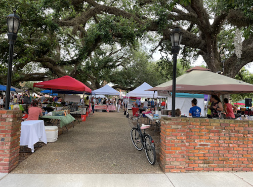 A bustling outdoor market with colorful tents, people browsing, and large trees providing shade.