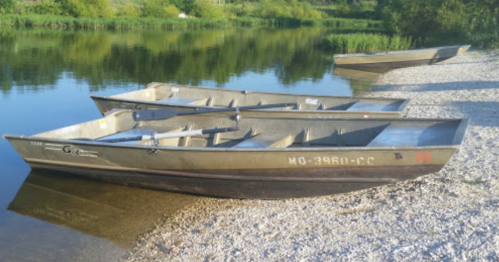 Three metal boats are moored on a pebbly shore beside a calm, reflective body of water surrounded by greenery.