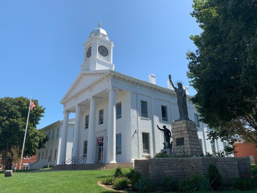 A white historic building with a clock tower, surrounded by greenery and a statue of a woman holding a torch.