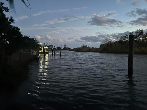 A calm waterway at dusk, reflecting light from a nearby structure, with trees and clouds in the background.