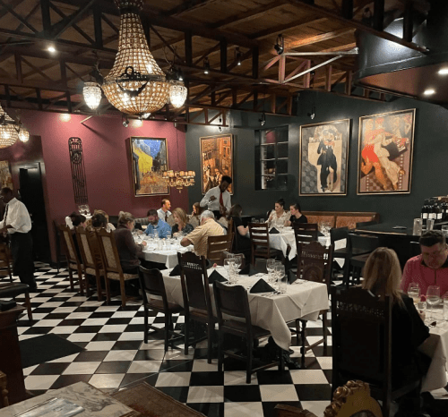 A lively restaurant interior with diners at tables, elegant decor, and large paintings on the walls.