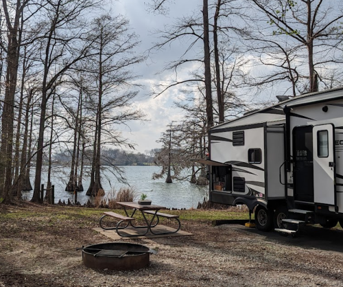 A camper parked by a lake surrounded by trees, with a picnic table and fire pit in the foreground.