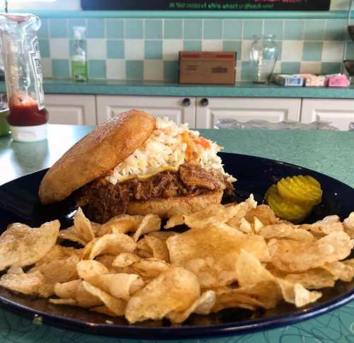 A pulled pork sandwich topped with coleslaw, served with a side of potato chips and pickles on a blue plate.
