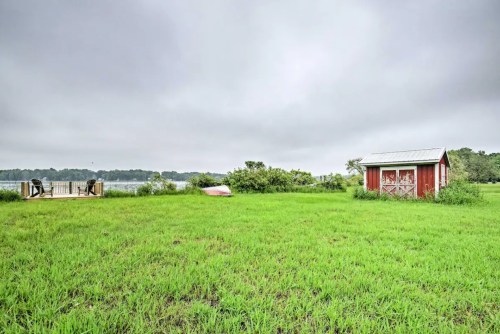 A grassy area with a red shed, a wooden deck by the water, and a cloudy sky in the background.