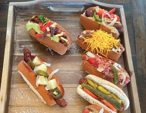 A wooden tray with six gourmet hot dogs, each topped with various colorful ingredients and garnishes.