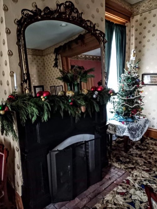 A decorated fireplace with greenery and ornaments, beside a small Christmas tree in a cozy, vintage room.