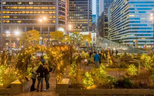A vibrant urban park at night, adorned with lights, featuring people mingling among greenery and city skyscrapers in the background.
