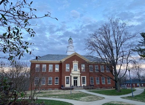 Historic brick building with a clock tower, surrounded by trees and a cloudy sky. Pathway leads to the entrance.