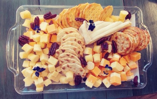 A platter of assorted cheeses, crackers, pecans, and decorative flowers, arranged artfully on a clear tray.