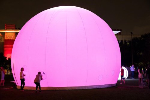 A large, glowing pink dome in a park at night, with people interacting around it.