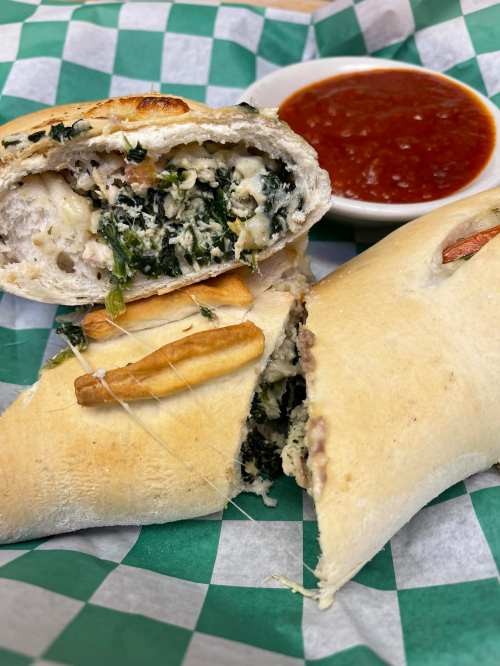 A close-up of a stuffed calzone with spinach and cheese, served with marinara sauce on a green checkered paper.