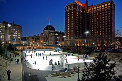 A bustling outdoor ice skating rink at night, surrounded by buildings and festive lights.