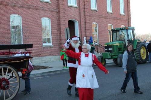 Santa and Mrs. Claus wave during a festive parade, with a tractor and holiday decorations in the background.