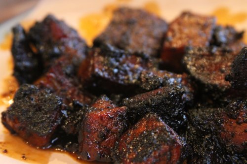 Close-up of charred, dark brown cubes of meat, glistening with sauce on a light plate.